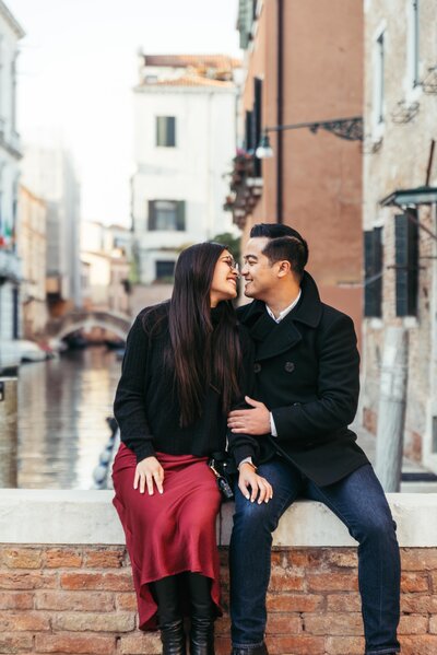 A couple sits on a wall while looking at each other lovingly, on the background a Venice Canal in Italy