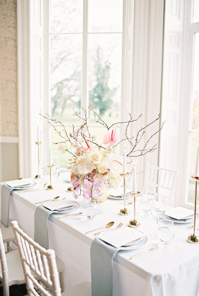 A white grey and pink wedding tablescape in front of large stately home windows and an opulent spring floral centrepiece.