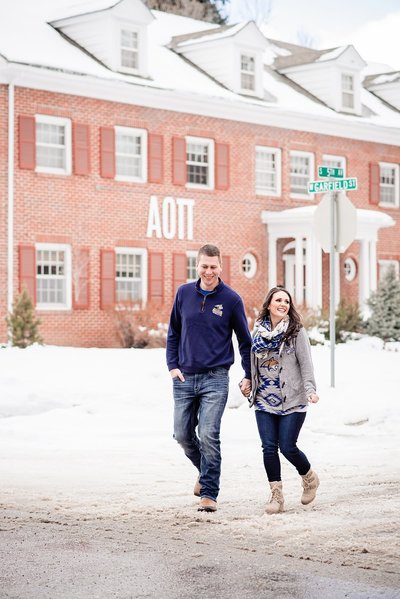 Couple wearing their Montana State bobcat attire and walking through snow covered street holding hands with the Alpha Omicron Pi Alpha Phi chapter house in the background