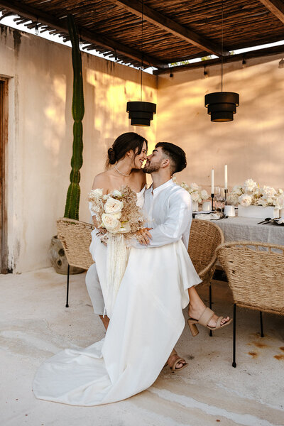 Bride sitting on groom’s lap at a modern Airbnb wedding table setup in Tulum, styled with neutral florals and candles.