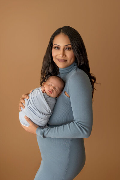 Smiling mother in a blue dress holding her swaddled newborn baby against a warm brown background.