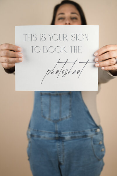 Brunette woman in overalls holding a sign that says this is your sign to book the photoshoot.