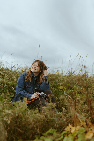 Alaska photographer smiling and sitting in a field of tall grass holding her camera