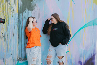 Two girls standing against a colorful wall smiling.