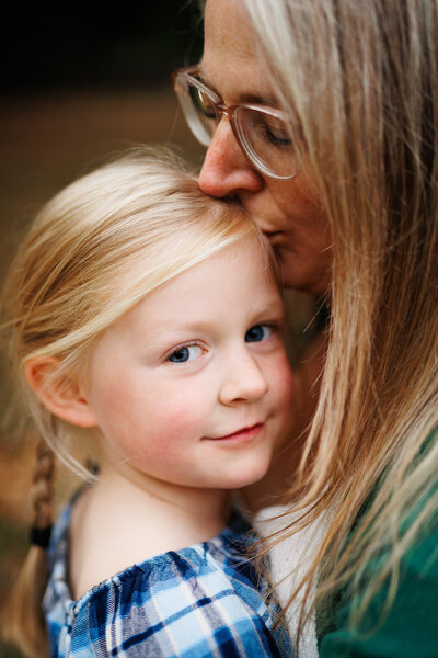 Tender moment between a mother and her young daughter, with the mother gently kissing the girl's head. The daughter, dressed in a blue plaid top, looks confidently at the camera.