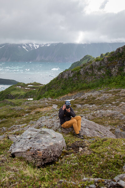 Photographer sitting on a rocky hillside in Alaska, taking a photo with an icy blue fjord and snow-capped mountains in the background under a cloudy sky.