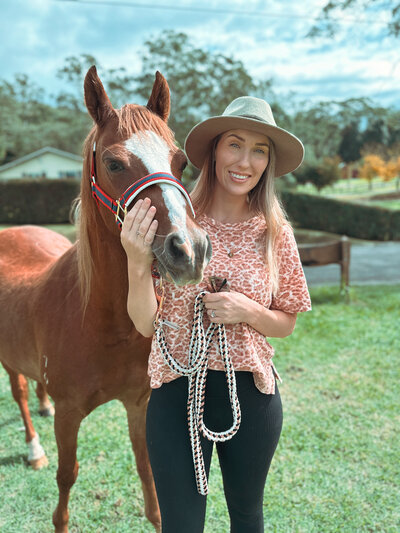 A women with a brown hat on, brown top and blue pants, sitting on a dark brown horse smiling. 