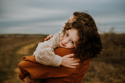 A toddler girl with curly hair laying her head on her mom's shoulder in a field of grass in Flower Mound, Texas