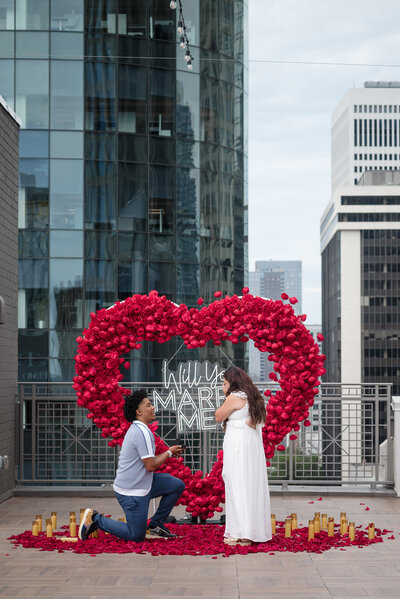Romantic Charlotte rooftop proposal with “Will You Marry Me?” sign and red rose heart, photographed by DeLong Photography, proposal photographer in Charlotte NC.