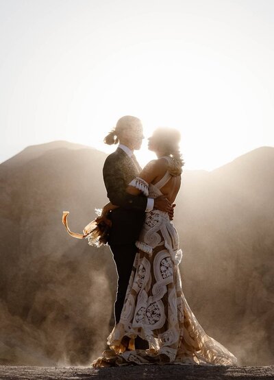 A couple shares a moment during their elopement, standing on a fallen tree in a golden meadow surrounded by tall pines, with a small rustic chapel and granite cliffs in the background in Yosemite National Park.