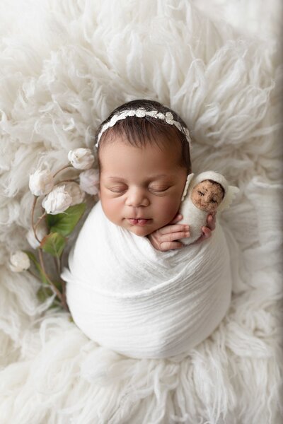 Studio newborn photoshoot in London, Ontario.  Newborn baby girls swaddled in white muslin blanket holding a felt doll against her cheek. She is wearing a delicate white headband on a white fluffy rug.