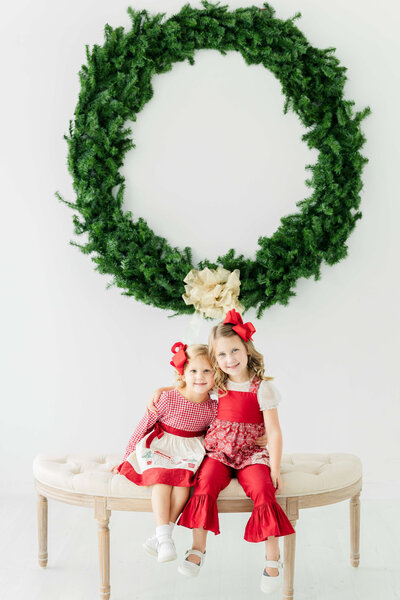 two girls sitting on bench in front of wreath for christmas mini session