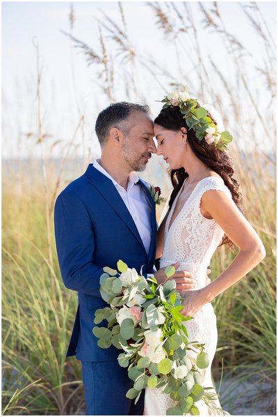 Groom embraces bride in flower crown in front of sea oats.