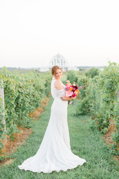 Bride holding bright pink floral bouquet stands amidst rows of vines at a vineyard