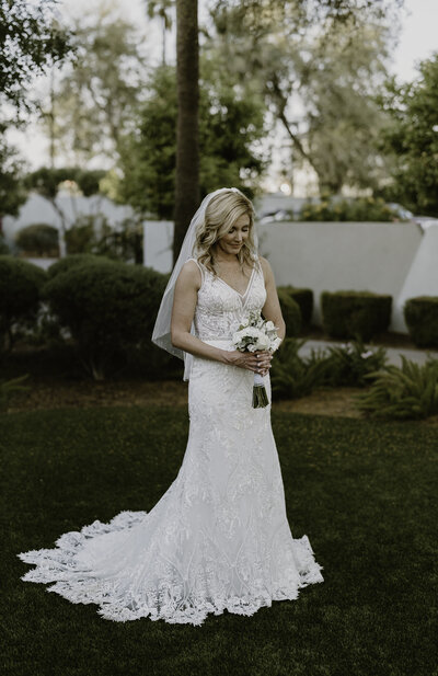 A bride in a detailed white lace wedding dress stands on a lush green lawn, looking down at her bouquet of white flowers. She has her blonde hair styled softly and a delicate veil falls down her back. Trees and a hint of a garden surround her, creating a serene and romantic setting