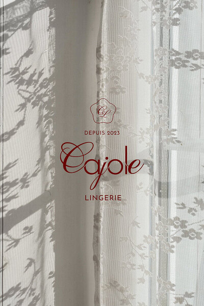 Maquette de présentation de l'identité visuelle de Cajole Lingerie, qui représente l'ambiance confortable et ensoleillée des matins au lit. L'image montre un rideau en dentelle et le logo vertical de la marque.