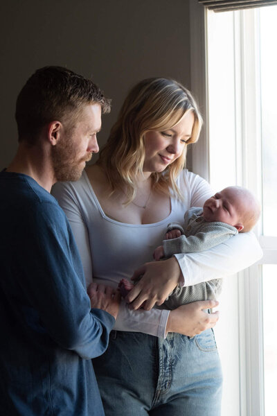 Happy new parents admire their sleeping newborn in mom's arms while standing in a window for a Colorado Springs Newborn Photographer