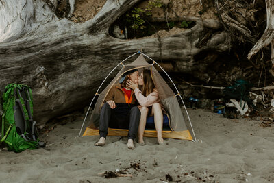 Hannah and Adam sitting in a tent on the beach and kissing