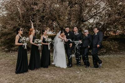 Bride and groom celebrate with their wedding party after the ceremony. The bride is popping a bottle of champagne. The entire wedding party is celebrating and laughing. Captured by top London, ON wedding photographer Ashlee Ellison.