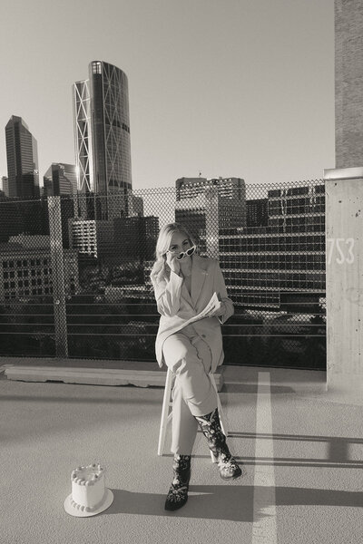Woman sitting on a rooftop with city buildings in the background, reading a newspaper outdoors.