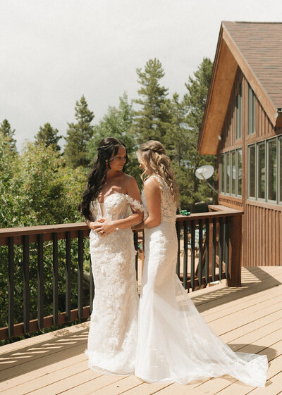 Two brides in lace gowns standing on a wooden deck with forest views
