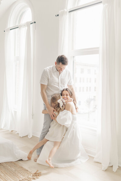 A family of three dressed in light colors hug each other during a photography session at a natural light studio in Kansas City. 