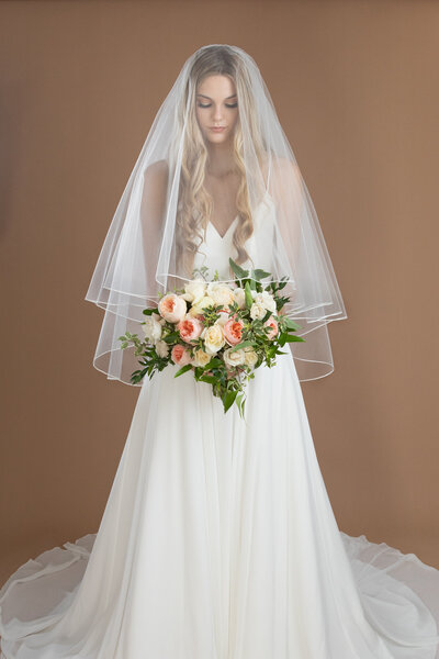 Bride wearing a fingertip length circle veil with blusher and small ribbon edge, and holding a white and blush bouquet