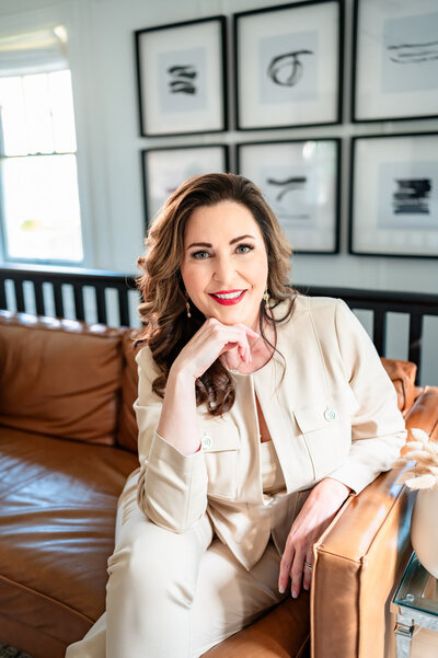 A woman with long brown hair and red lipstick is sitting on a tan leather couch, wearing a light-colored suit, smiling, and resting her head on her hand. Black and white framed art is on the wall behind her.