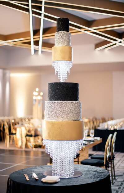 A tall, elegant wedding cake with six tiers, alternating black, silver glitter, and gold layers. Crystal-like decorations hang between each tier, creating a cascading chandelier effect. Background features a modern venue with seating.