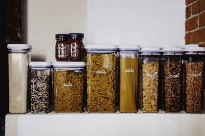 Organized pantry shelf with clear labeled containers and jar of pasta and grains, set against a white wall and accent wall of a warm brick wall.
