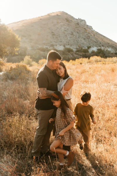 A mother and father embracing while their two young kids run around them.