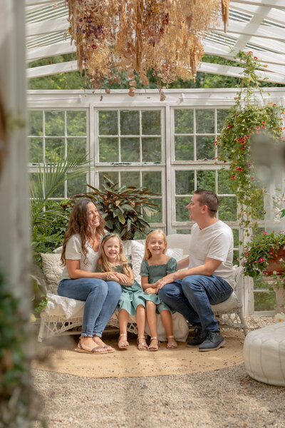 family of four with two young girls sit on white wicker couch in beautiful greenhouse decorated with lots of greenery at rustic wild arrow in brooklyn michigan