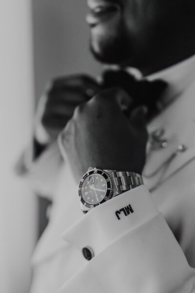 a close up photo of a groom straightening his bow tie with his watch and monogram of his initials in focus