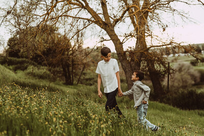 Two young boys holding hands, looking at each other, walking up a green meadow in Fresno, CA 