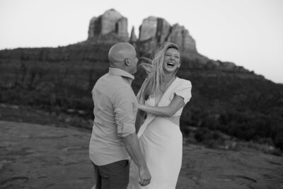 a black and white image of a couple, the woman's face visible and laughing, the back of the man's head is only visible. Sedona red rocks in the background
