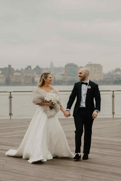 Bride and groom holding hands while walking on a waterfront boardwalk with a city skyline in the background on a cloudy day.