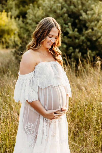 stunning mama to be in a white lace dress holding her baby bump and looking down as she thinks about her journey to motherhood captured by Door County Photographer Ashley Kalbus