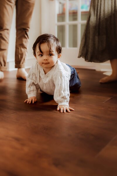 A baby in a white sweater and jeans crawls on a wooden floor, while three adults stand nearby.