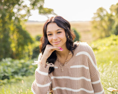 A high school senior girl smiling at the camera, with her hand under her chin, smiling at the camera, with a light tan and white striped shirt, pink nail polish, and has dark brown hair and eyes