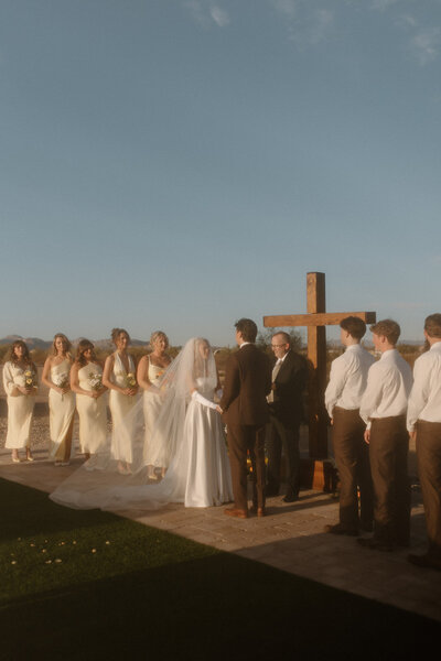 a photo that looks like film of a wedding ceremony with a cross at the alter. the bridesmaids are in pale yellow dress and the bride is dress in a classically elegant old money style dress with elbow length gloves