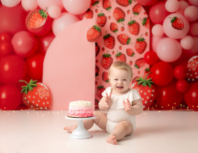 one year old girl in white onesie sitting next to cake with strawberry themed backdrop