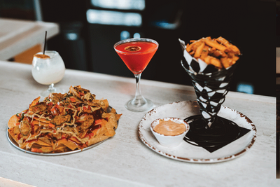 Juicy bacon cheeseburger topped with a cherry tomato and pickle skewer, served alongside crispy golden fries in a tin and a refreshing cold beer.
