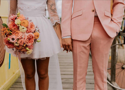 Gros plan sur les tenues des mariés se tenant par la main. Robe courte blanche pour elle et costume rose pour lui. Immortalisé par Laura, photographe en Vendée.