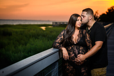 A loving maternity portrait of a couple on a boardwalk at sunset; the man in black gently kisses the forehead of the pregnant woman in a beautiful black lace gown, with marshland and water in the background
