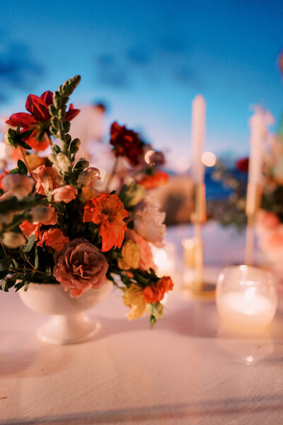 A close-up of a colorful floral centerpiece with pink, red, and orange flowers, surrounded by candlelight on a wedding reception table at dusk