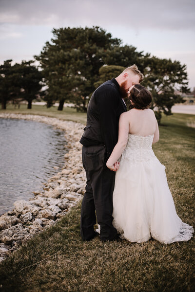 A couple stands hand-in-hand near a pond, with trees in the background, sharing an intimate moment.