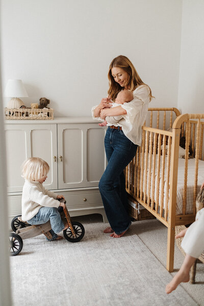 mum in nursery holding baby, while toddler riding tricycle 