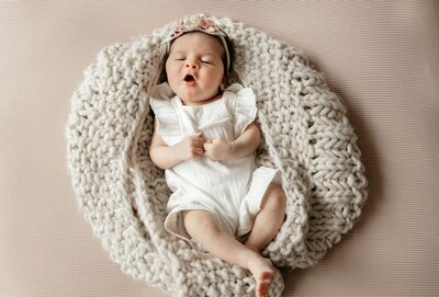Newborn baby girl yawning while lying on a chunky cream knit blanket, wearing a white ruffled outfit and a floral headband by Ruaa Hamdan