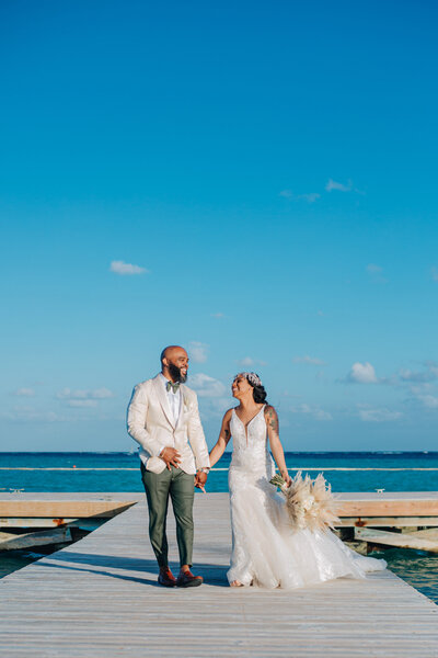 A bride and groom walk hand in hand along a wooden dock by the ocean. The groom wears a white blazer and green pants, while the bride wears a detailed lace gown and holds a bouquet of pampas grass.
