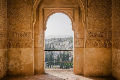A stone archway with intricate carvings frames a picturesque view of a distant hillside town, reminiscent of scenes curated by a European travel specialist, with scattered buildings and lush greenery under a hazy sky.